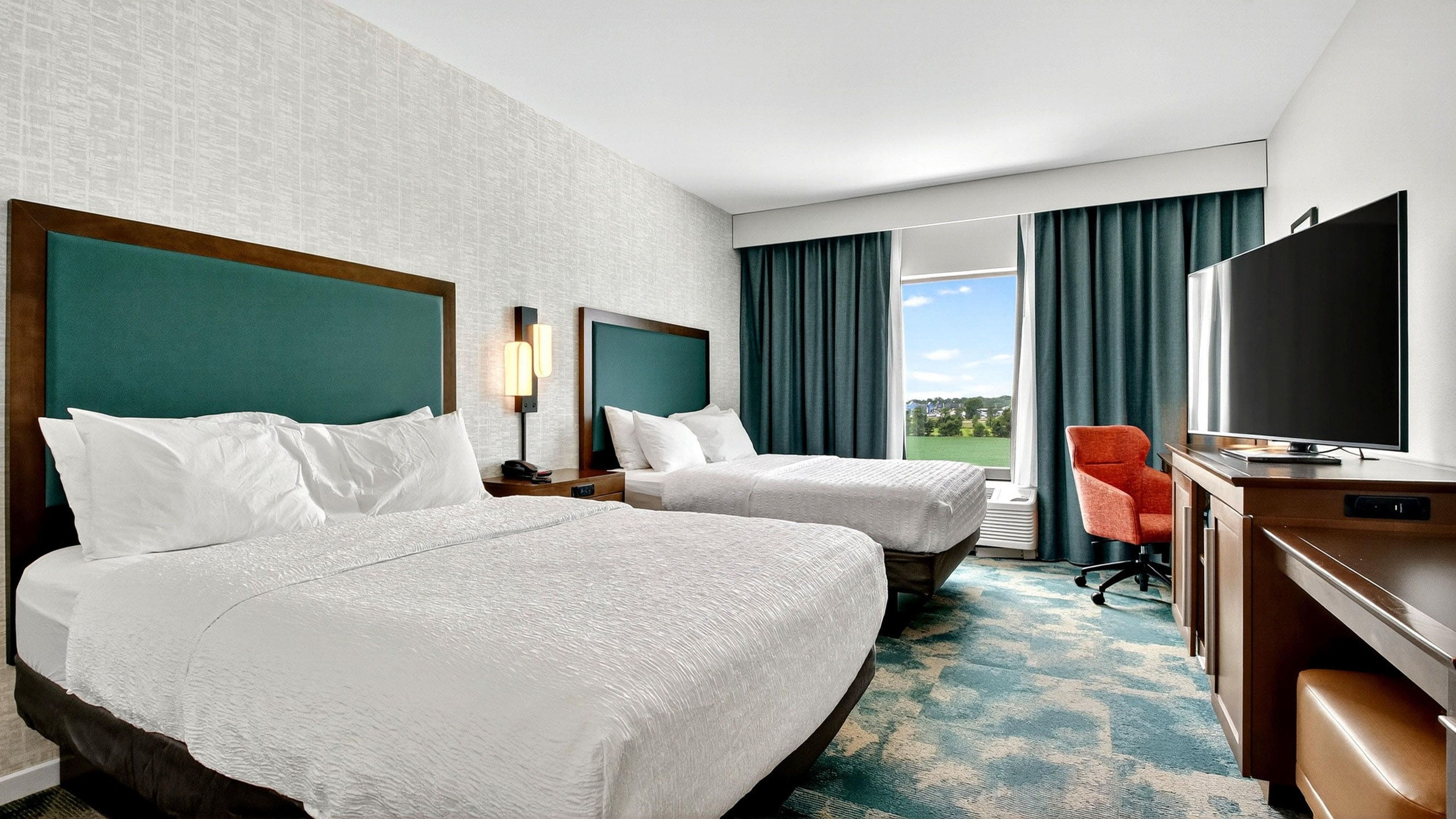 A hotel room with two beds, a desk with a flat-screen TV, a chair, and a view of the outdoors.