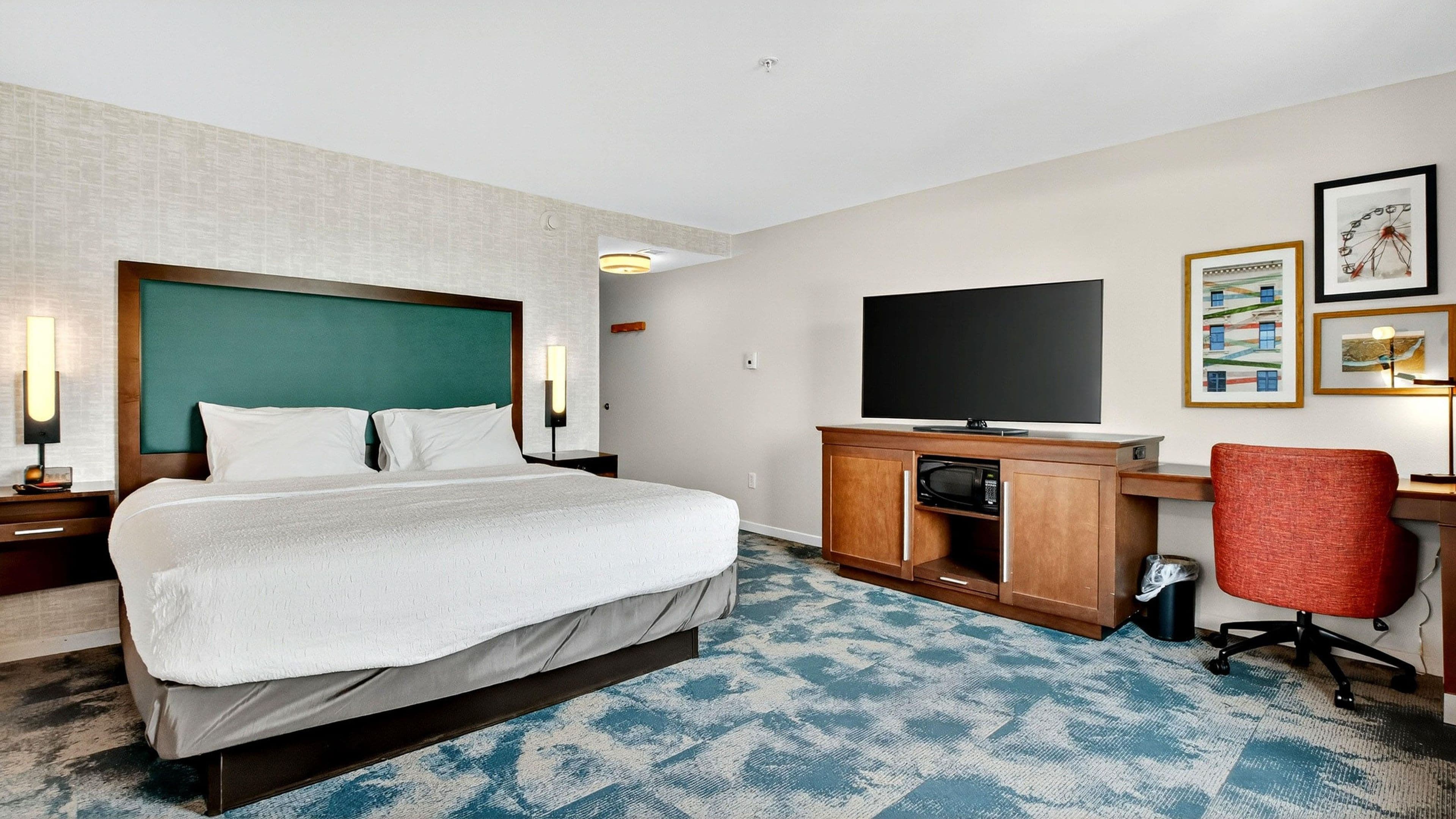 A hotel room with a bed, a desk with a flat-screen TV, a red chair, and a patterned carpet.
