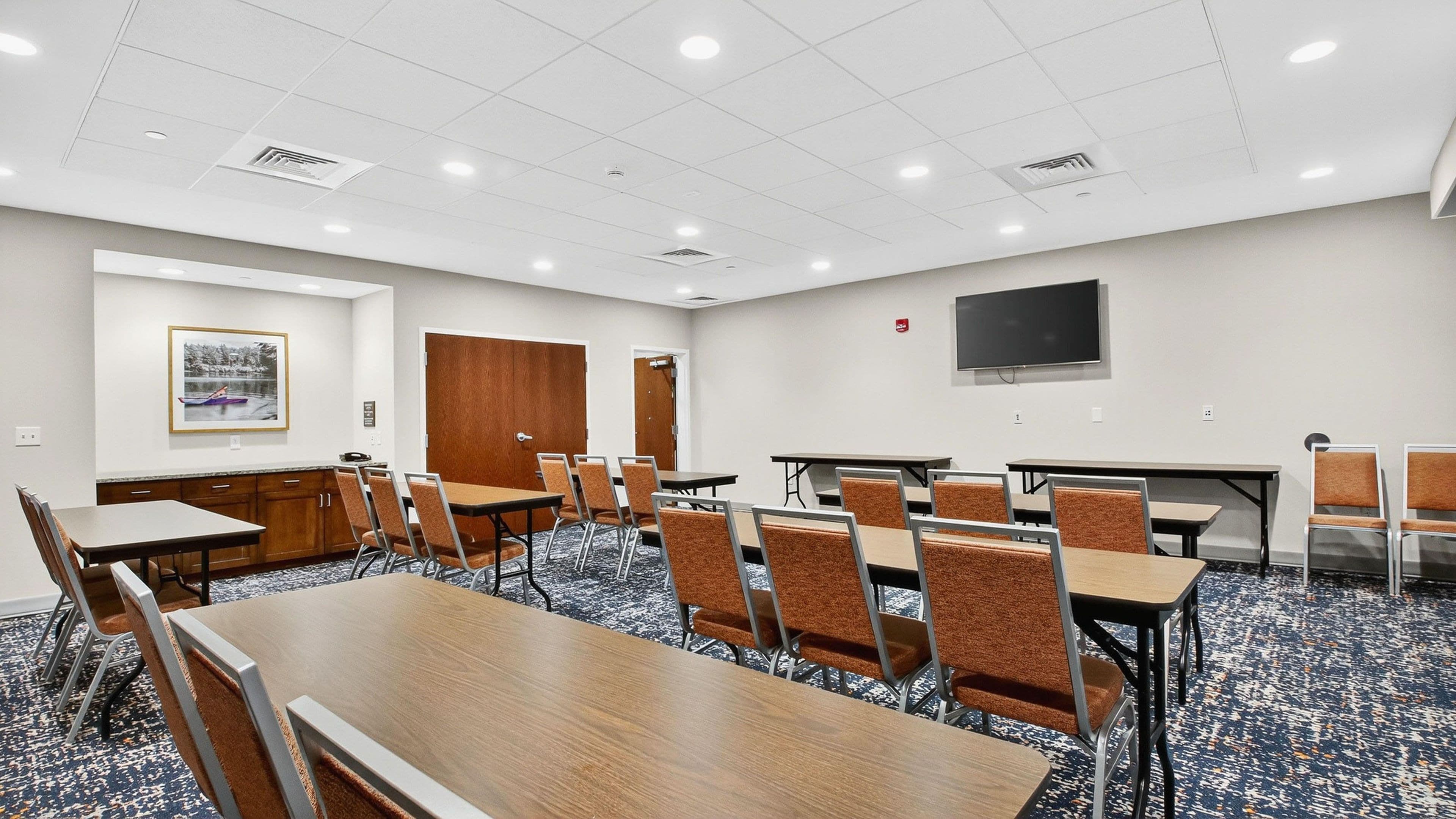 A conference room with multiple tables and chairs, a door, a framed picture on the wall, and a flat-screen TV mounted on the wall.