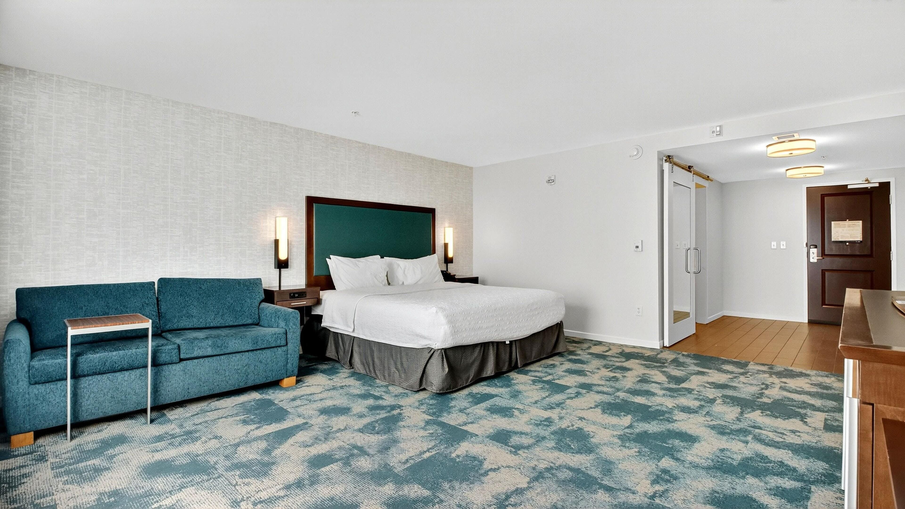 A hotel room with a bed, a blue sofa, a small table, and a nightstand with a lamp.