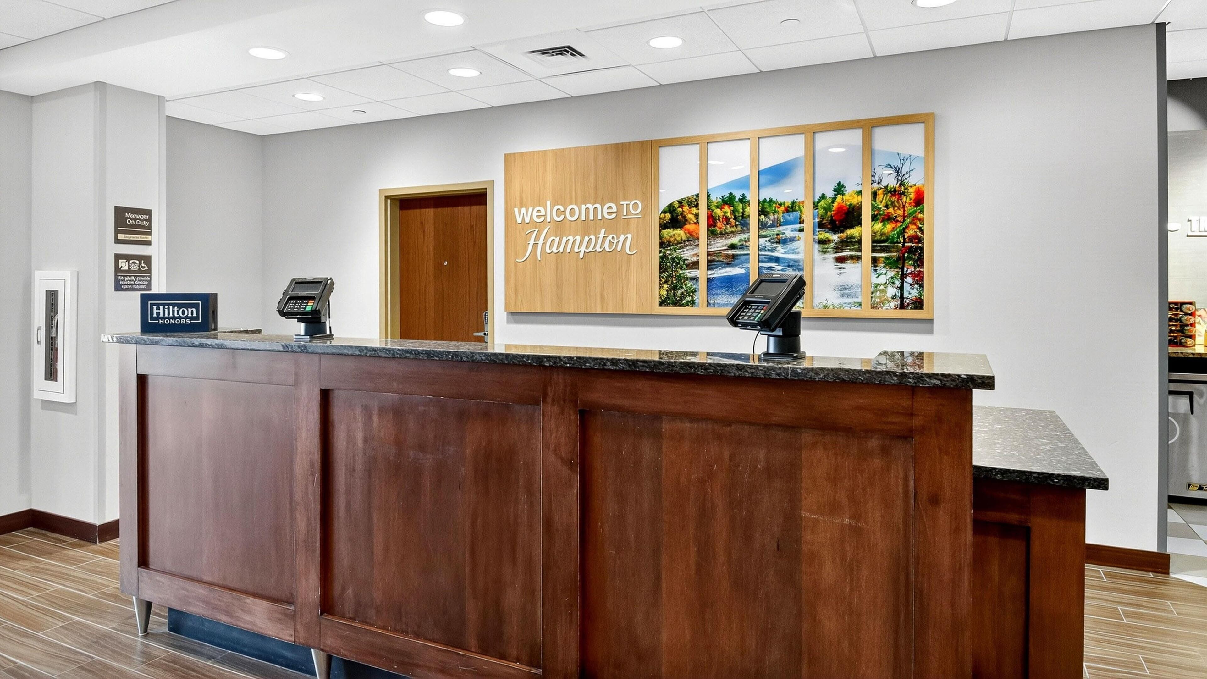 A reception desk with a wooden finish, a 'Welcome to Hampton' sign, and a 'Hilton Honors' badge.