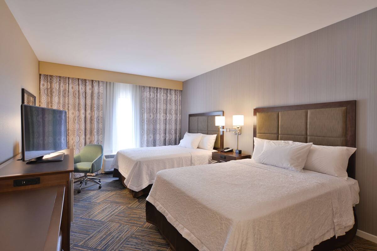 A hotel room with two beds, a desk, a chair, and a TV.