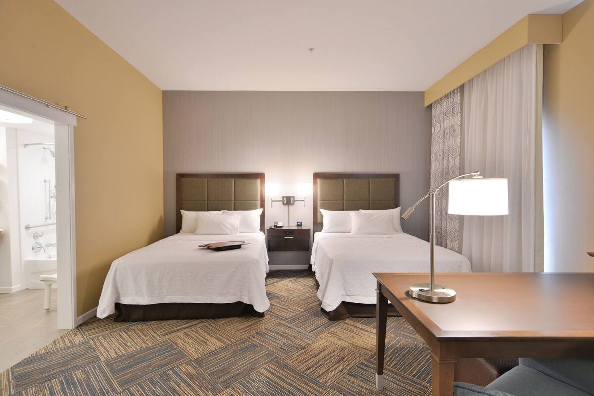 A hotel room with two beds, a desk, a chair, a lamp, and a bathroom visible through an open door.