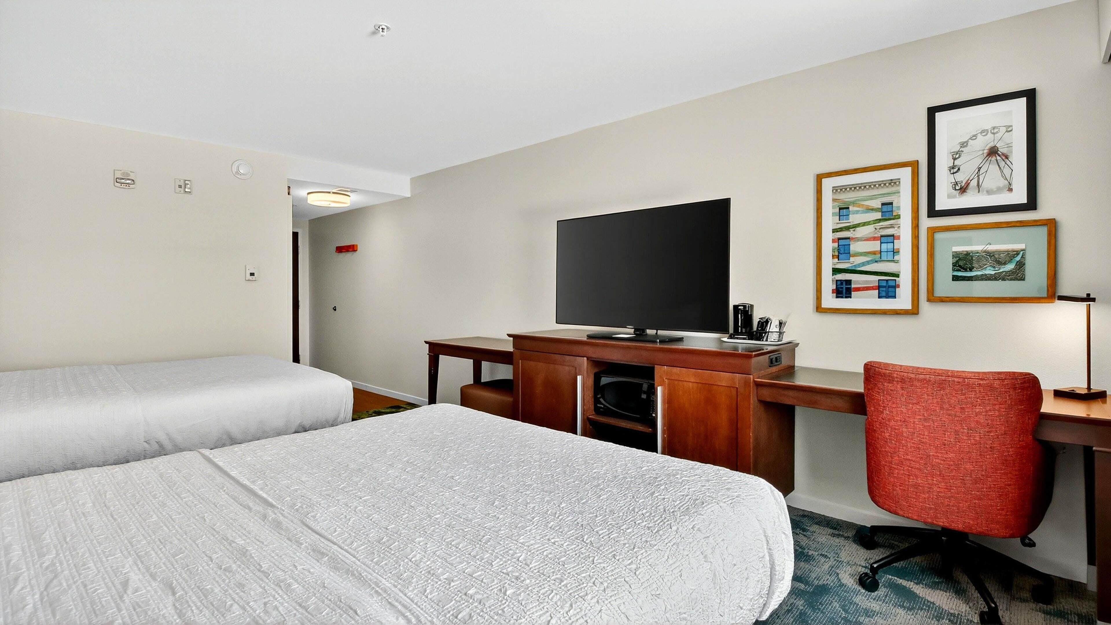 A hotel room with two beds, a desk with a flat-screen TV, a red office chair, and framed pictures on the wall.
