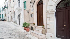 A narrow street lined with white buildings, each with a brown door and green shutters. There are potted plants on the steps of one building. - B&B Casa Cimino (Monopoli)