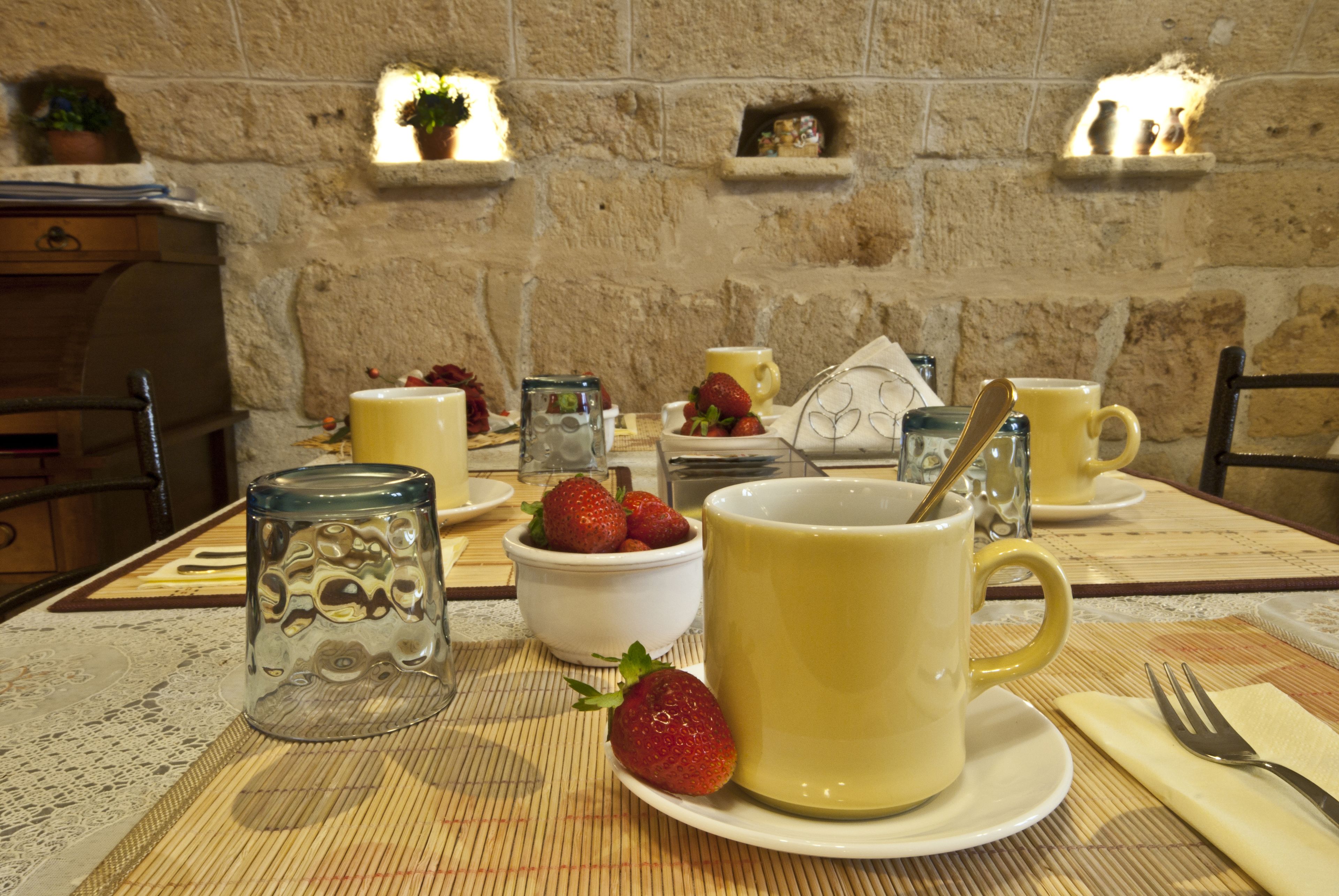 A breakfast setting with a stone wall, a table with a placemat, and a variety of breakfast items including a bowl of strawberries, a glass jar, and yellow cups.