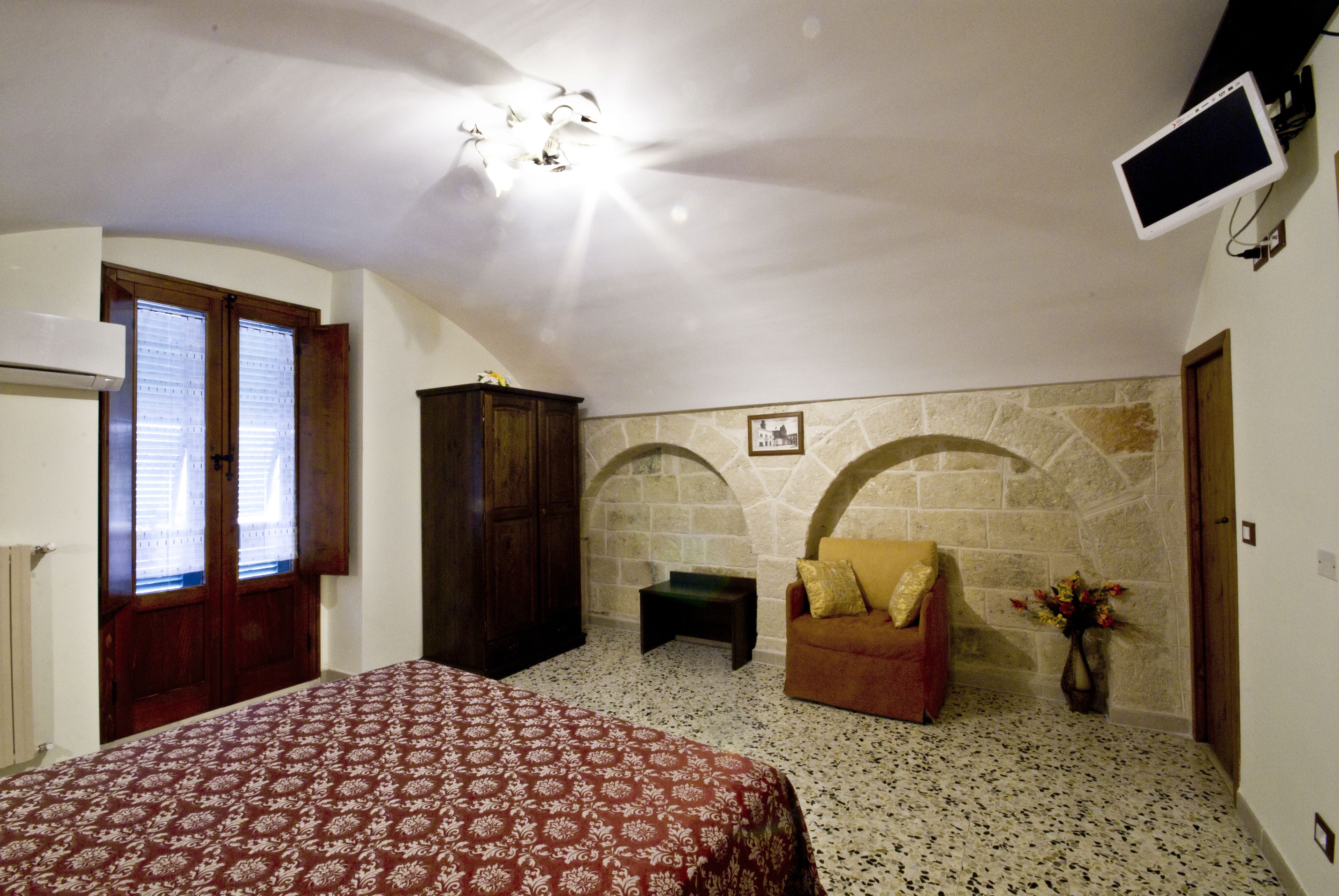 Un dormitorio con cama, armario de madera, mesita pequeña, sofá, pared de piedra y televisor montado.