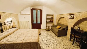 A bedroom with a bed, a sofa, a small table, and a chair. There is a window with blinds, a radiator, and a stone wall. - B&B Casa Cimino (Monopoli)