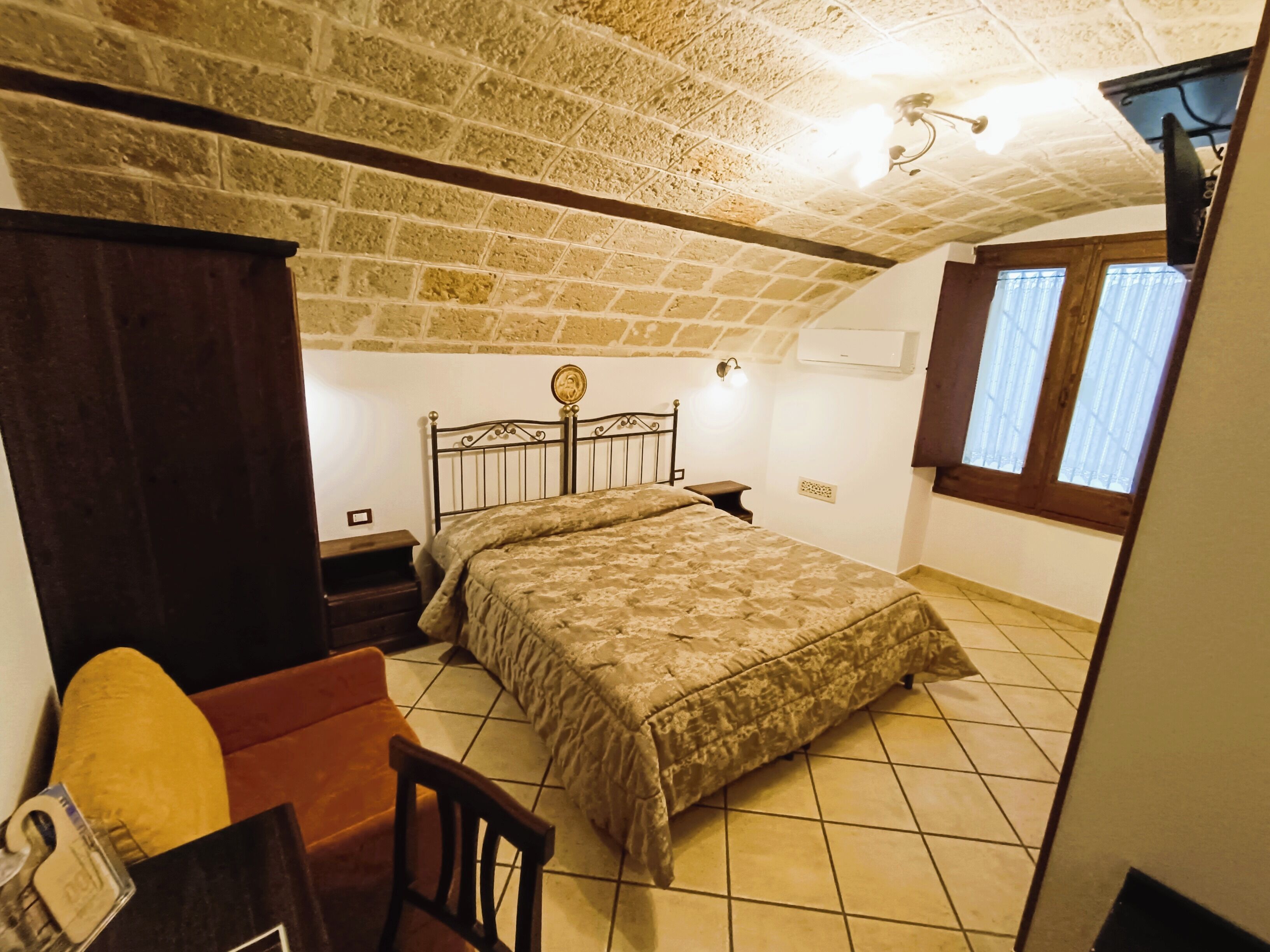 A bedroom with a stone ceiling, a bed with a headboard, a nightstand, a chair, and a window with curtains.