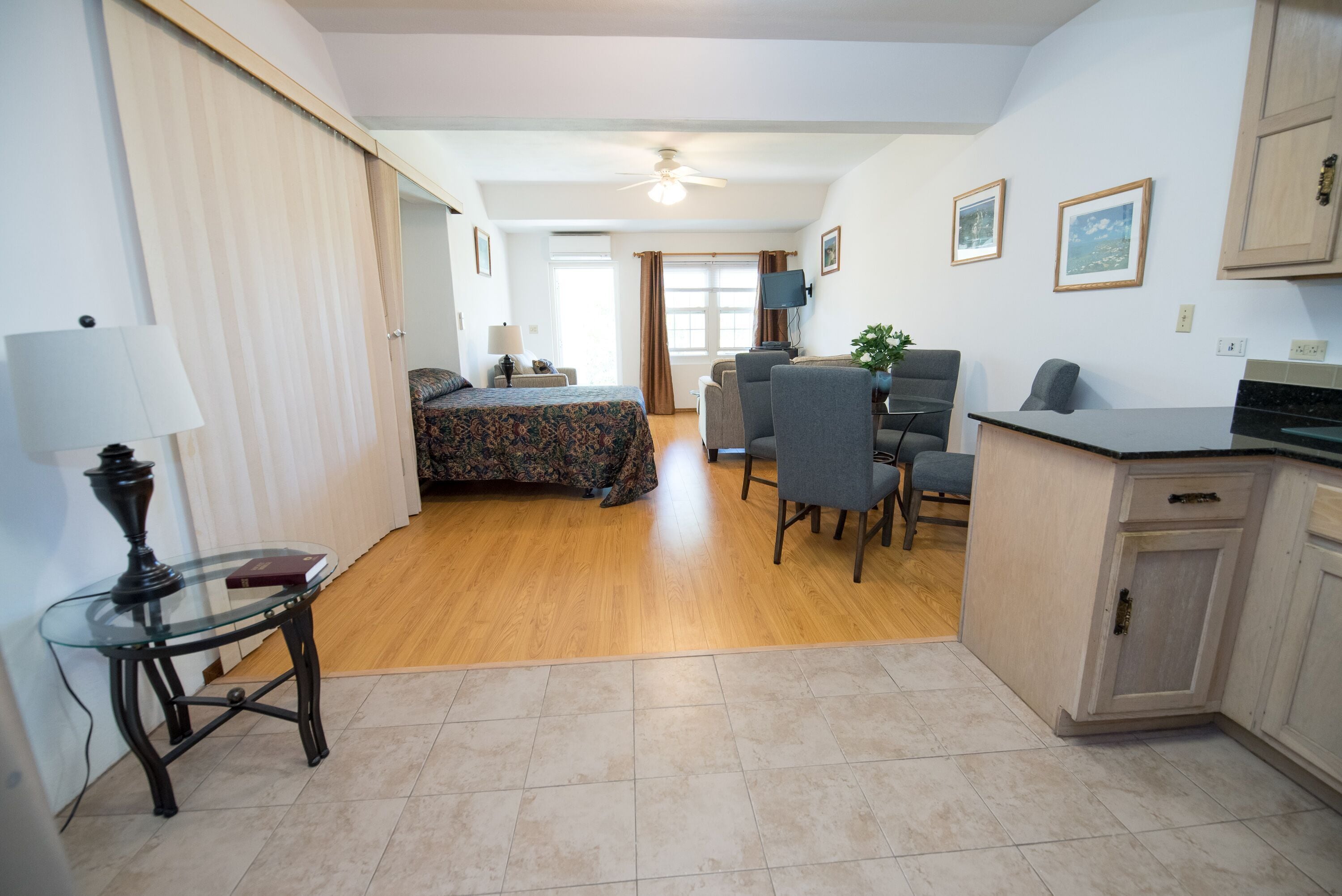 A hotel room with a bed, a desk, a chair, a small table with a lamp, and a kitchenette area.