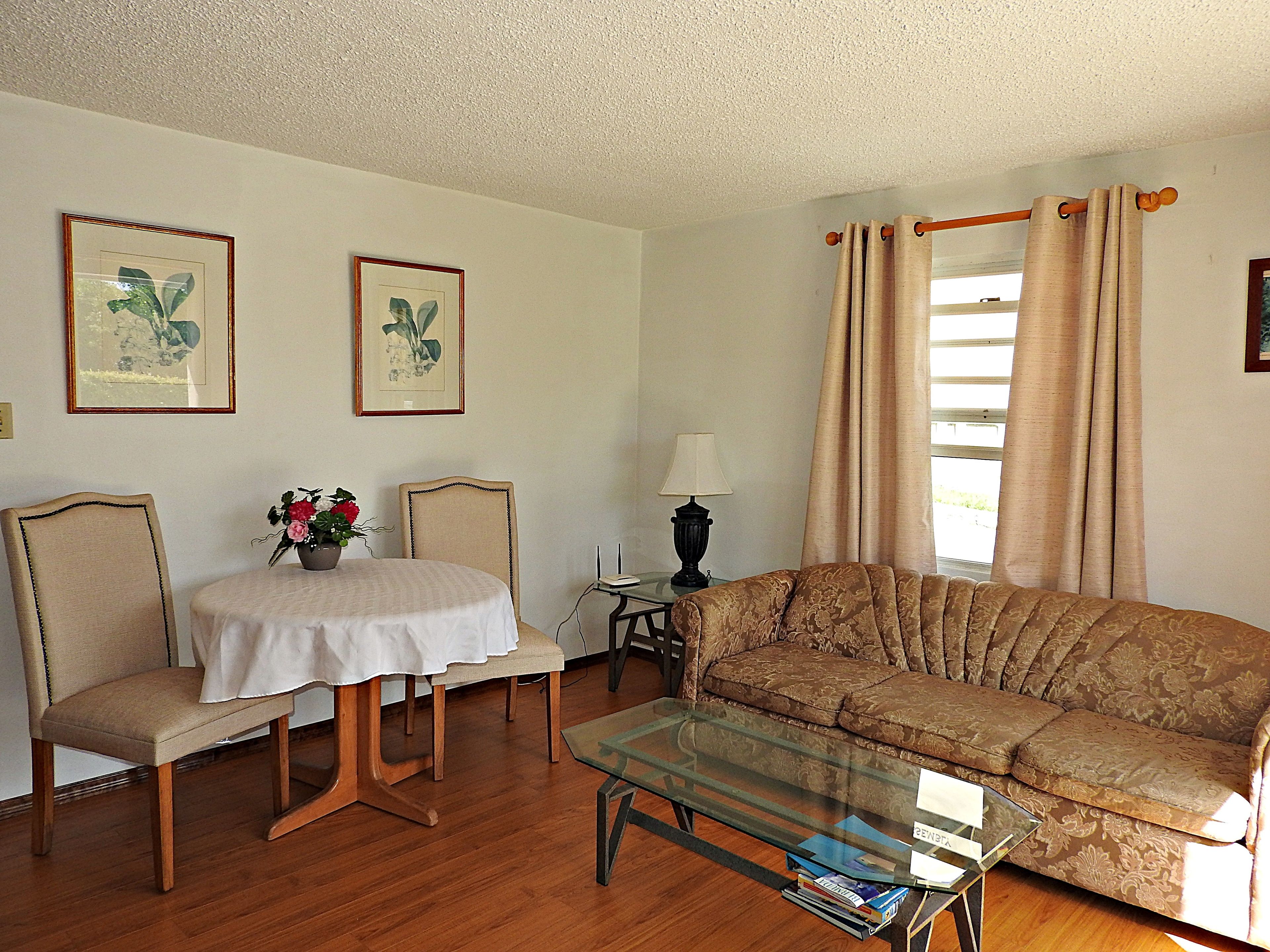 A living room with a sofa, two chairs, a glass coffee table, a small table with a lamp, and two framed pictures on the wall.