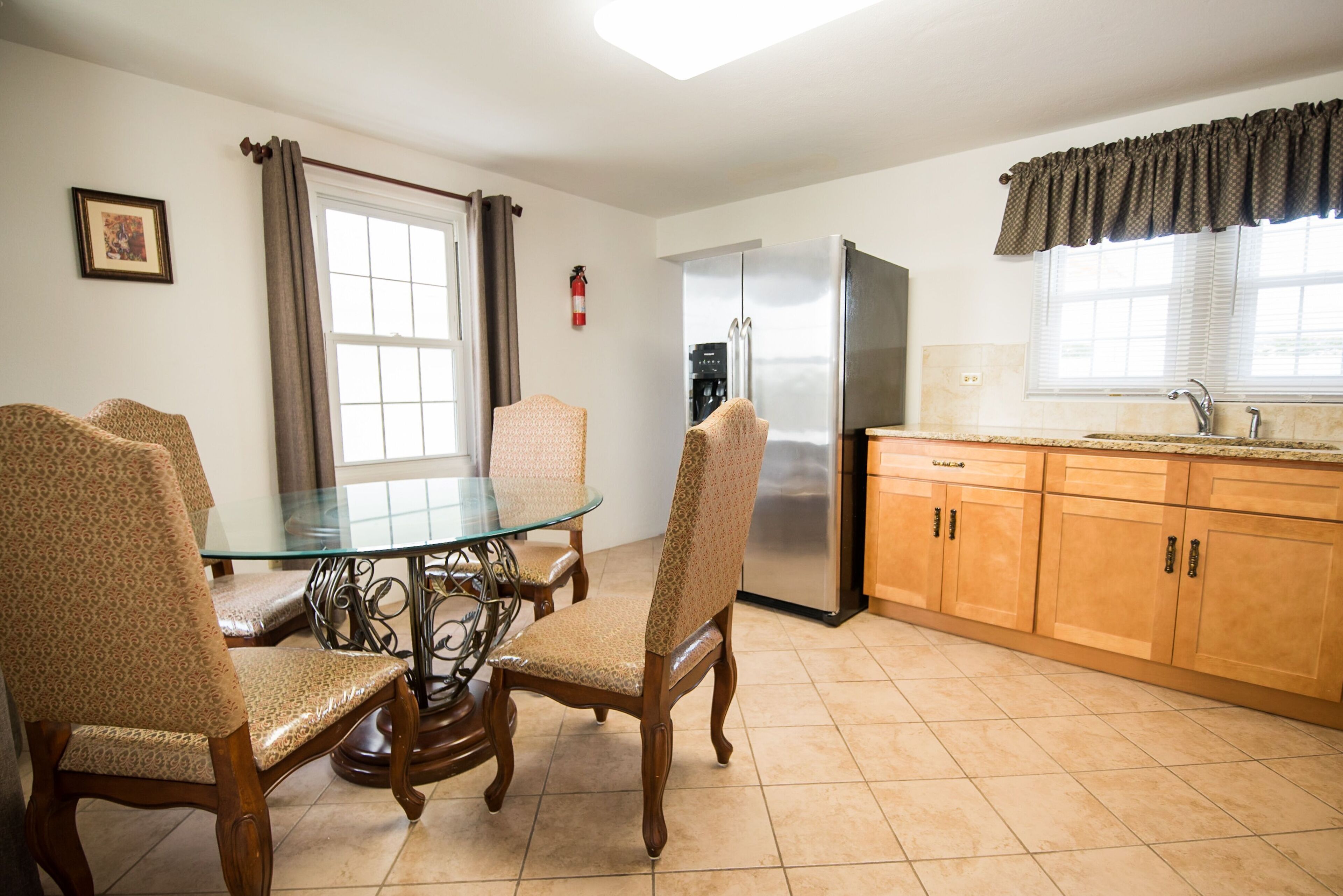 A kitchen with a glass-top dining table, wooden chairs, a stainless steel refrigerator, wooden cabinets, and a window with curtains.