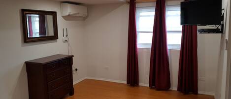 A room with wooden flooring, a dark wooden dresser, a wall-mounted air conditioner, a flat-screen TV mounted on the wall, and a window with red curtains.