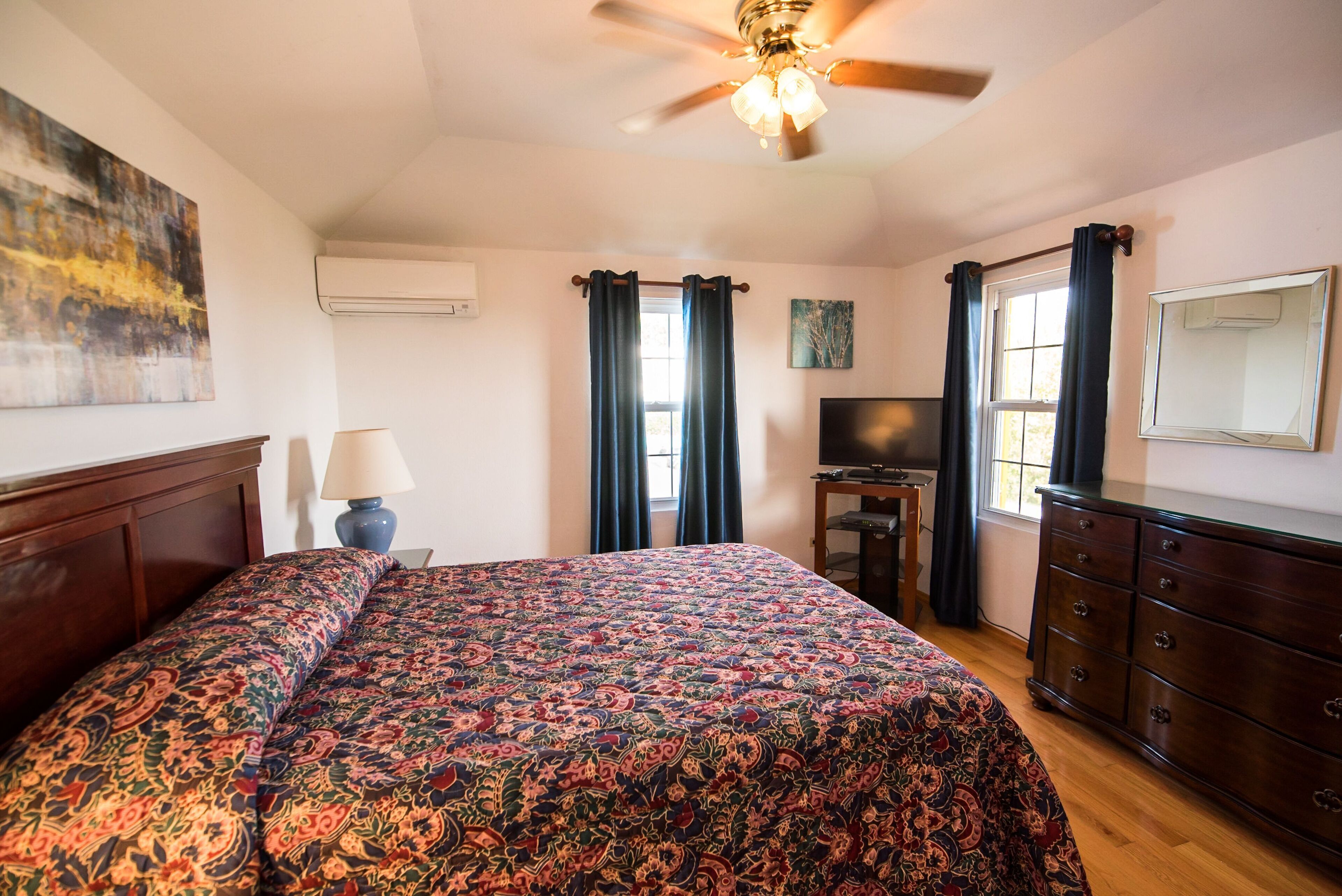 Une chambre à coucher avec un grand lit, un ventilateur de plafond, une télévision, une commode et une fenêtre avec des rideaux.