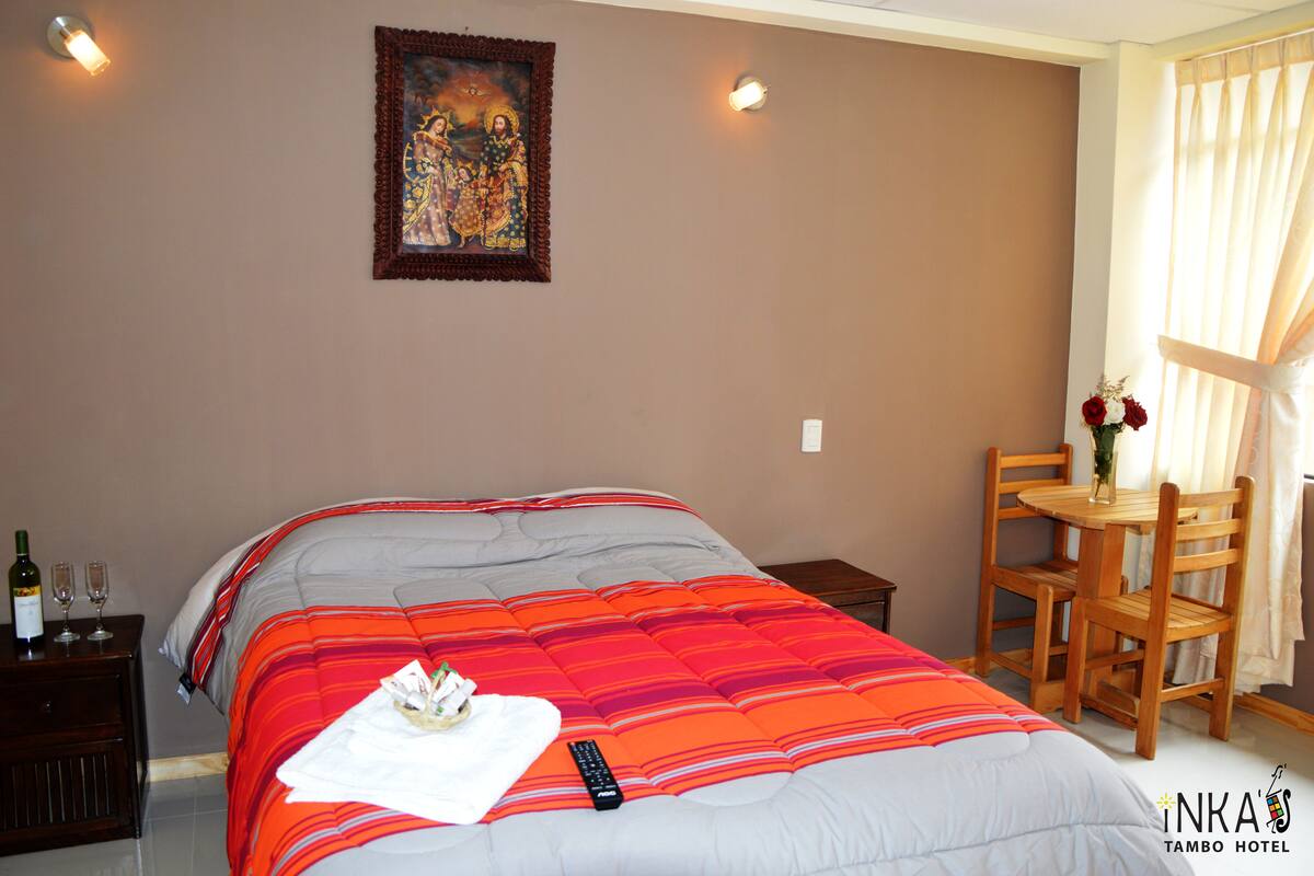 Una habitación de hotel con una cama, una mesita con sillas, un cuadro en la pared y una botella de vino en la mesita de noche.