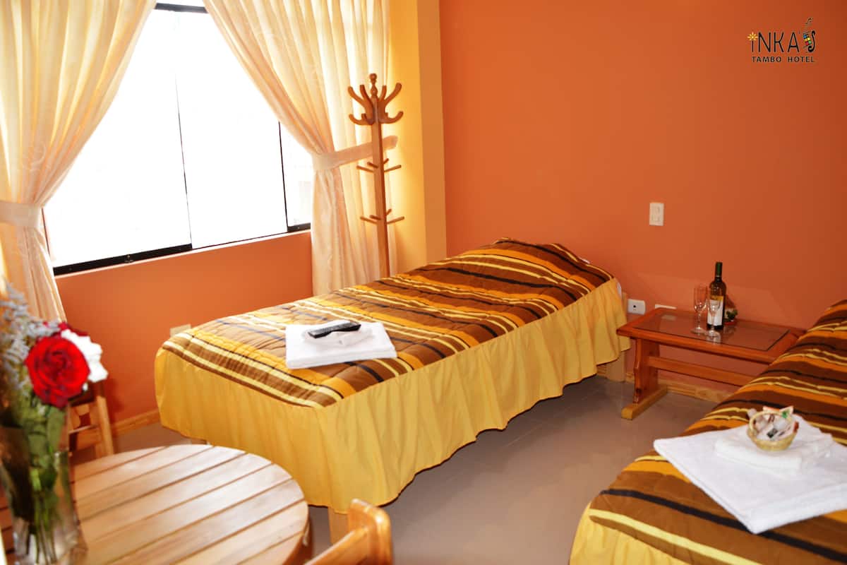 Habitación de hotel con cama, mesita de noche con botella y vasos, una silla y una ventana con cortinas.