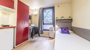 A compact hotel room with a kitchenette, a bed with a purple pillow, a desk with a laptop, and a window with a view of bare trees.