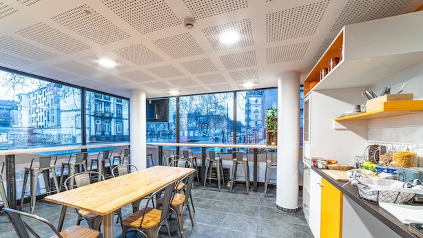 A modern cafe with a wooden table and metal chairs, large windows with city views, and a kitchen area with shelves and utensils.