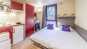 A compact hotel room with a kitchenette, a bed with two pillows, a window with a view, and a small desk with a computer.