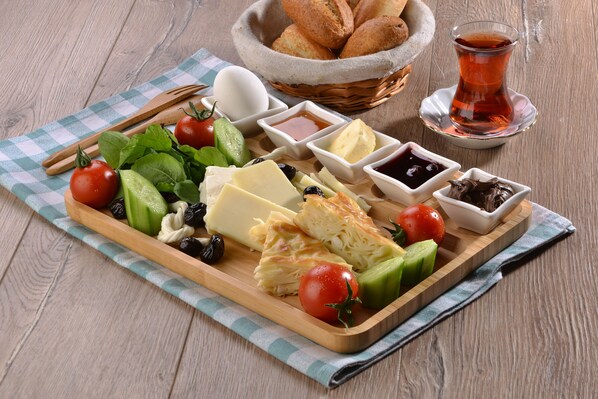 Un plateau en bois contenant divers fromages, des olives, des tomates, des concombres et un bol de pain.