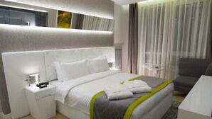 Una habitación de hotel moderna con una cama grande, mesitas de noche, un escritorio y vistas a la ciudad a través de cortinas translúcidas.