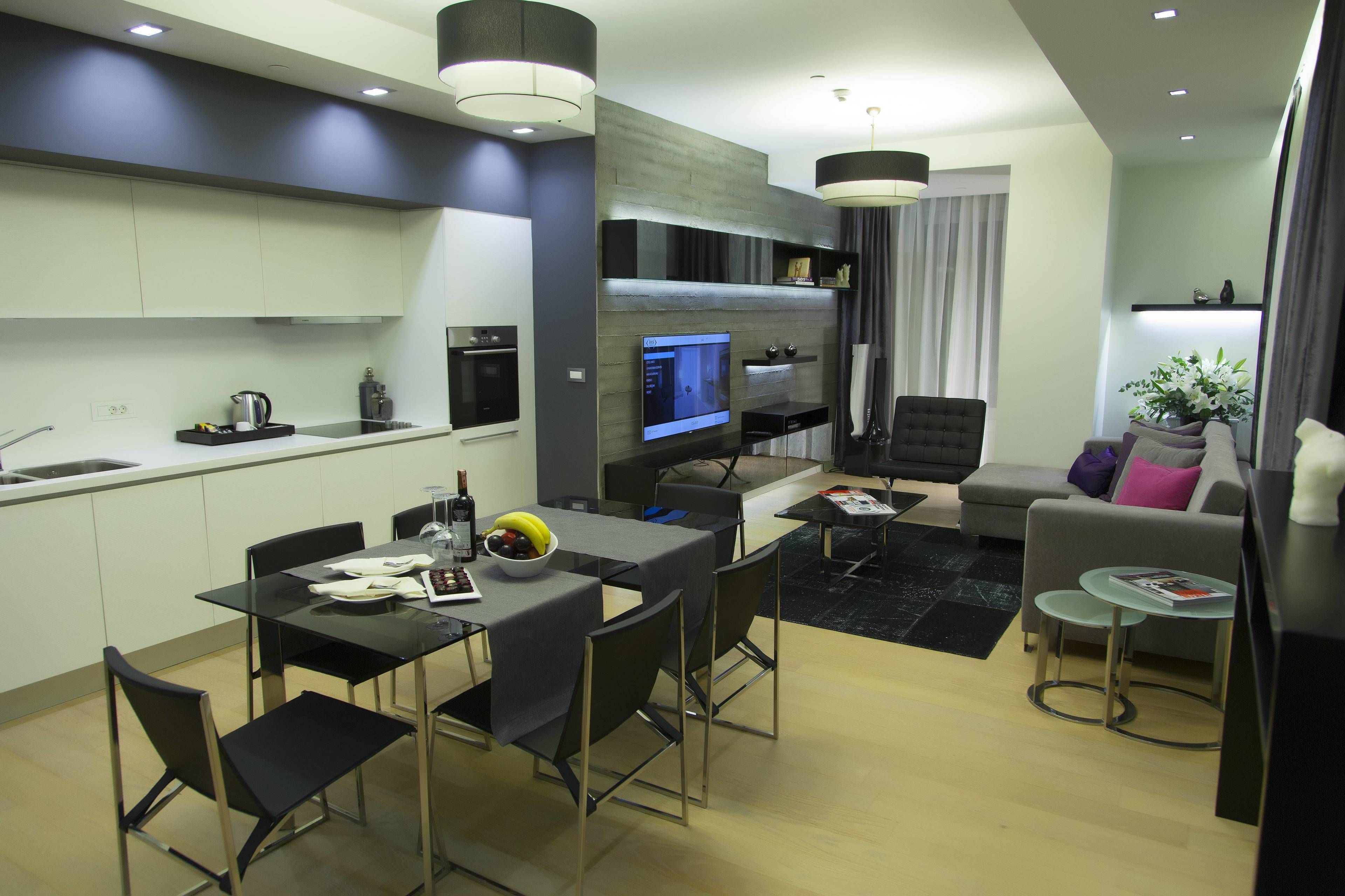 A modern kitchen with a built-in oven, a dining area with a glass table and chairs, a living room with a sofa and a coffee table, and a TV mounted on the wall.