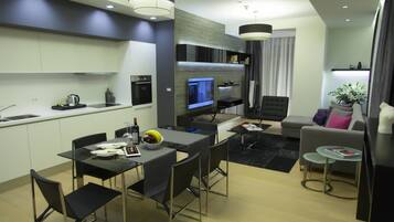 A modern kitchen with a built-in oven, a dining area with a glass table and chairs, a living room with a sofa and a coffee table, and a TV mounted on the wall.
