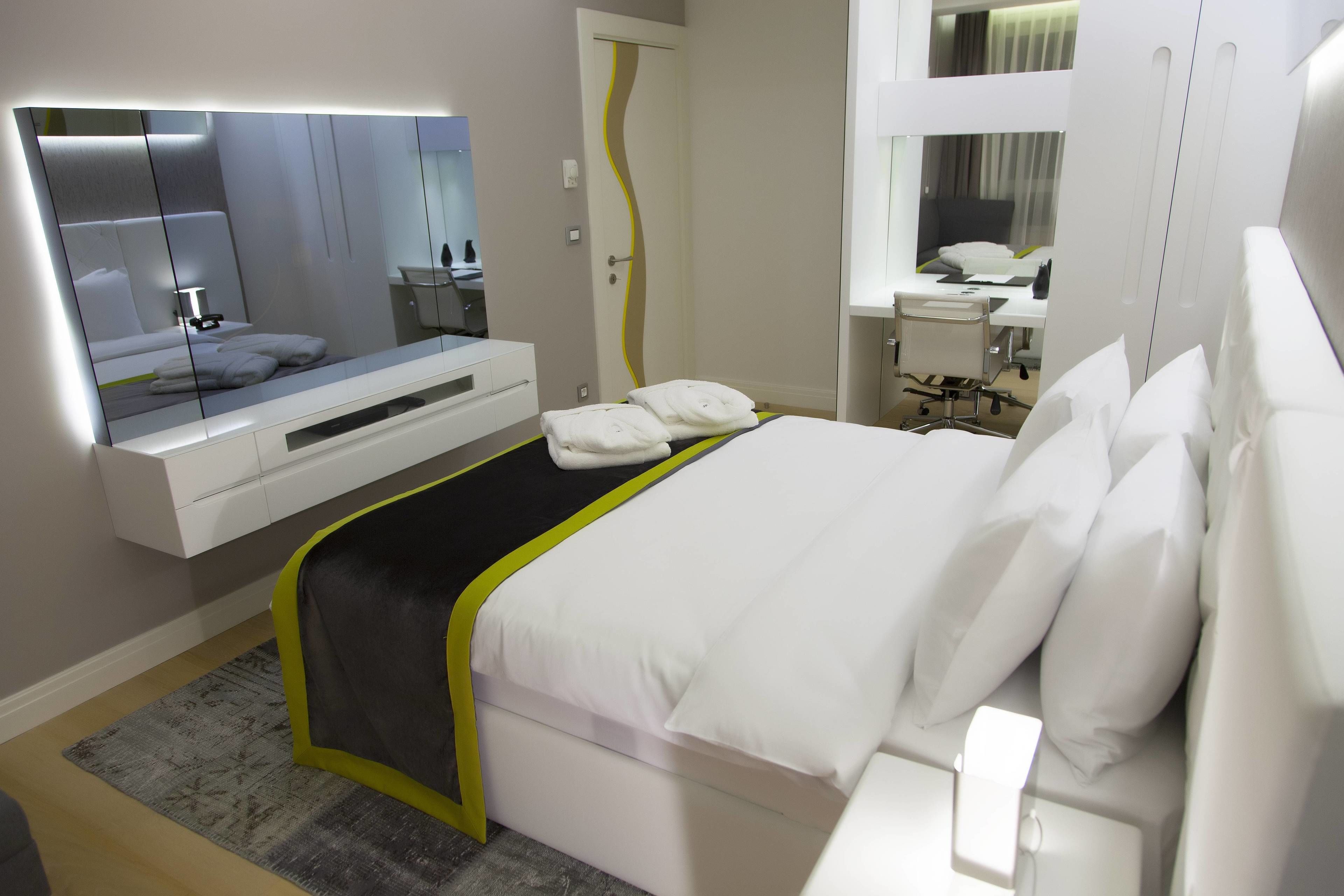 A modern hotel room with a large bed, a bedside table, a mirror, and a bathroom with a sink and toilet.
