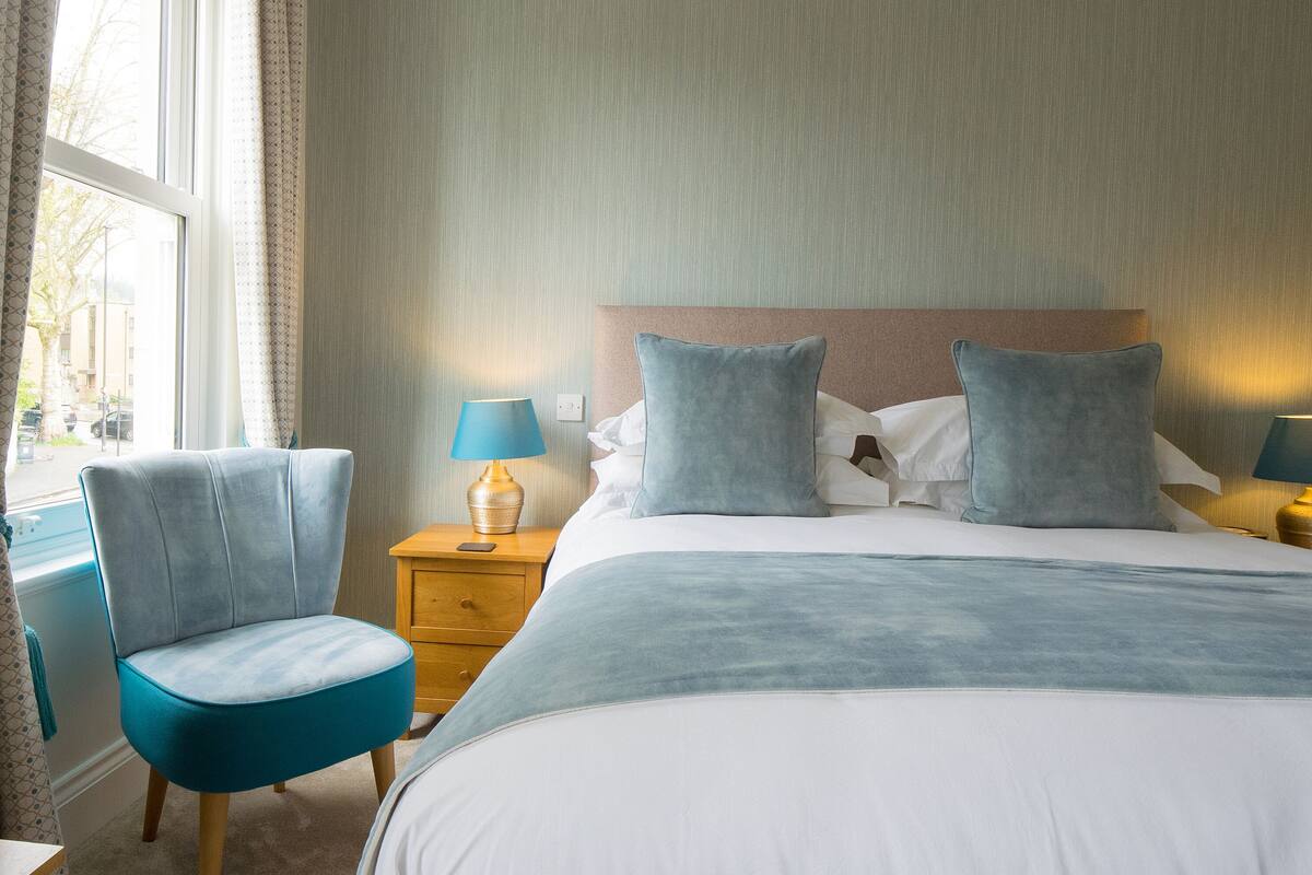 A hotel room with a bed, two bedside lamps, a chair, a nightstand, and a window with curtains.