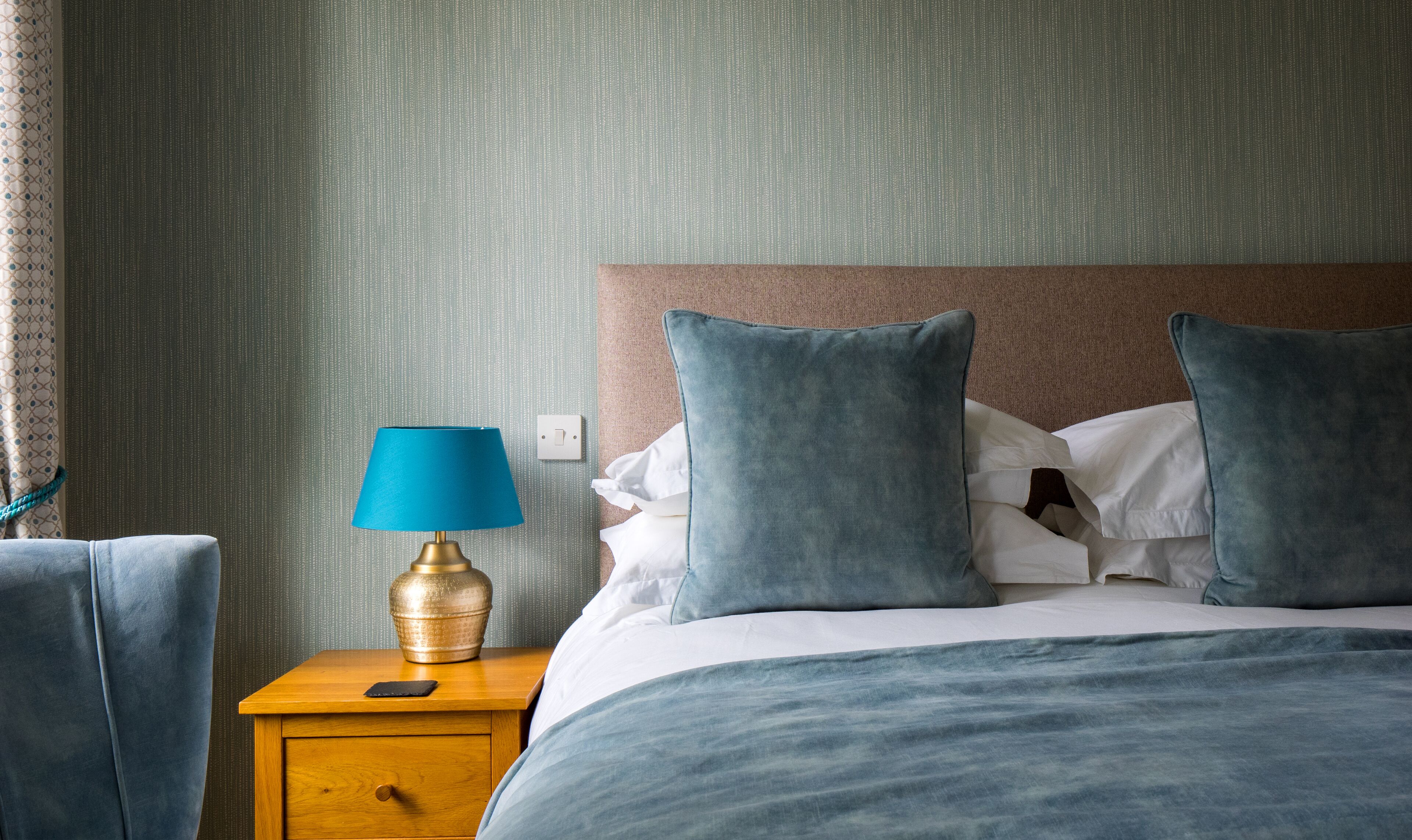 A neatly made bed with blue bedding, a wooden nightstand with a lamp, and a patterned wall.