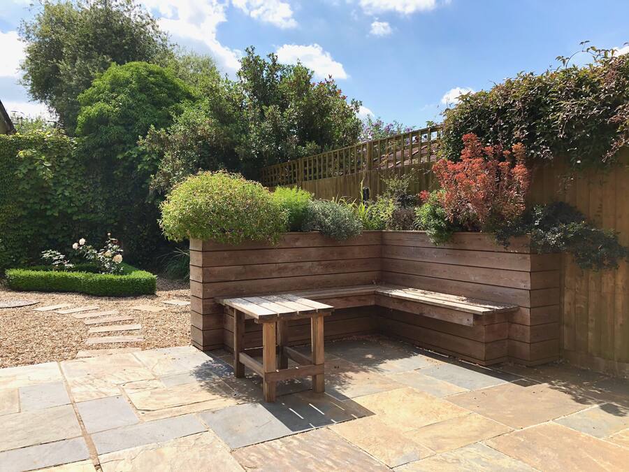 A wooden bench in a garden with stone tiles, surrounded by various plants and a wooden fence.
