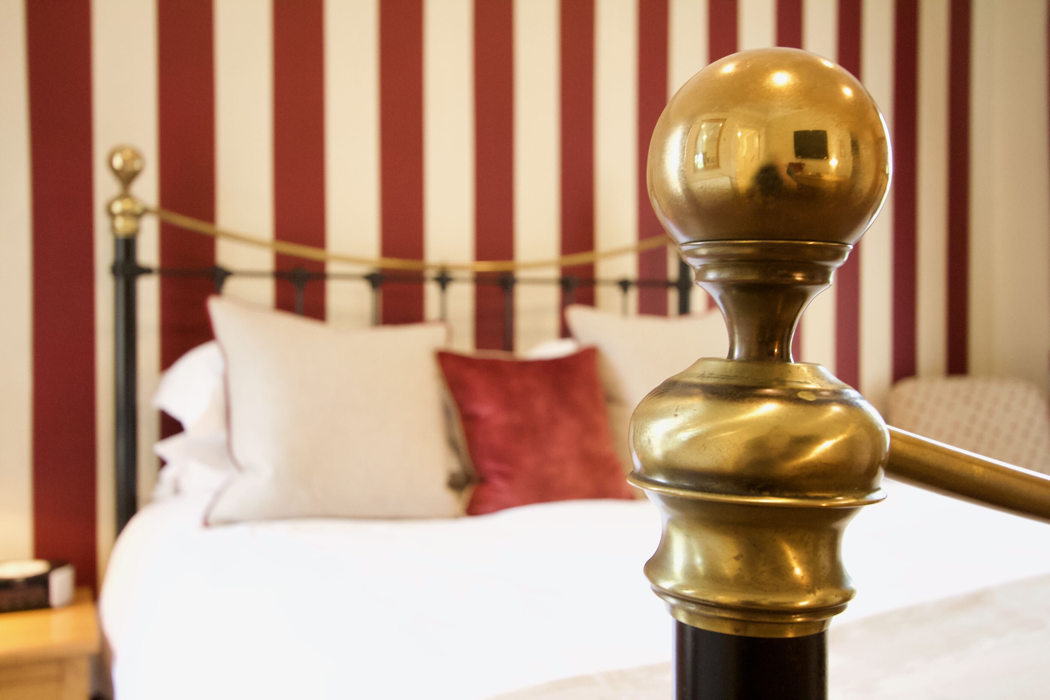 Una cama con almohadas blancas y rojas, un cabecero de bronce y una pared a rayas al fondo.