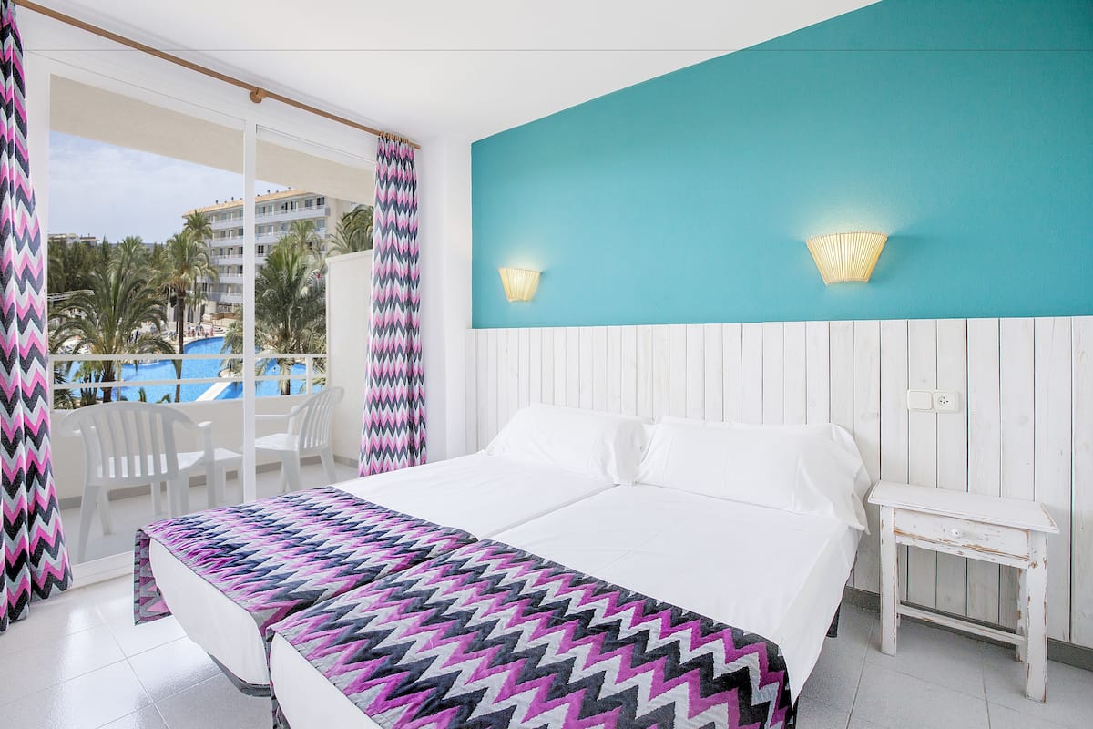 A hotel room with a bed, a small table, a balcony with chairs, and a view of a pool and buildings.