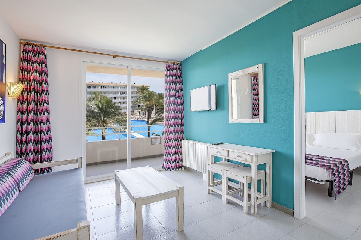 A hotel room with a bed, a desk, a chair, a table, and a view of a pool.