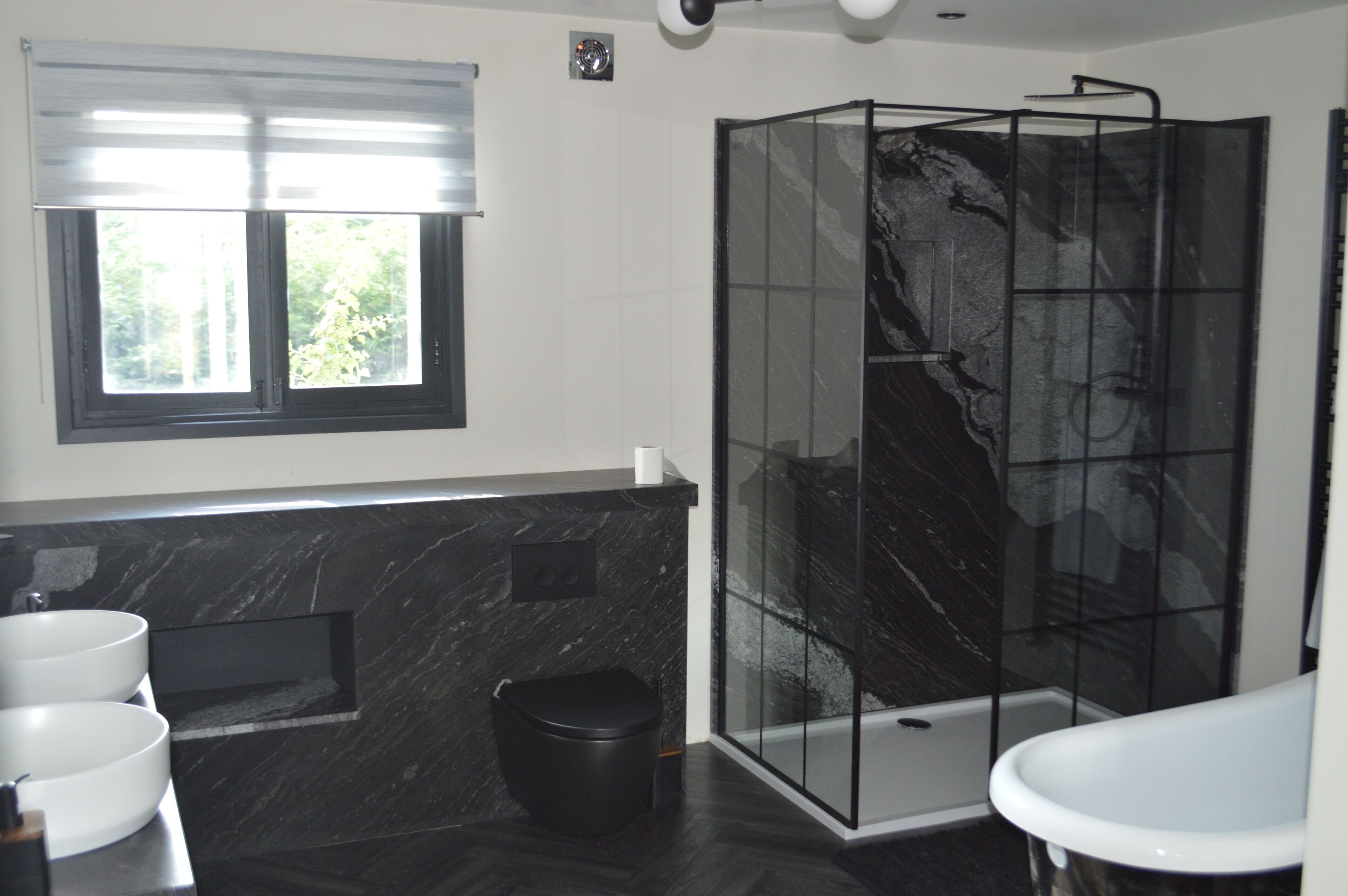 A modern bathroom with a black marble countertop, a glass shower enclosure, and a window with blinds.