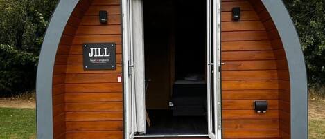 Una cabaña de madera con un letrero que dice "JILL" y una puerta abierta hacia el interior. Hay dos sillas afuera sobre una zona cubierta de pasto.