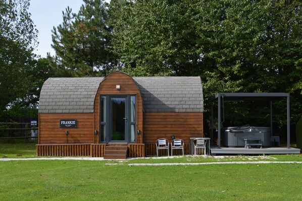 A small, single-story cabin with a wooden exterior and a gray roof, named "FRANKIE." It features a front door, a small porch, and a hot tub area with a covered seating space. - Heart House NEC (Coventry)