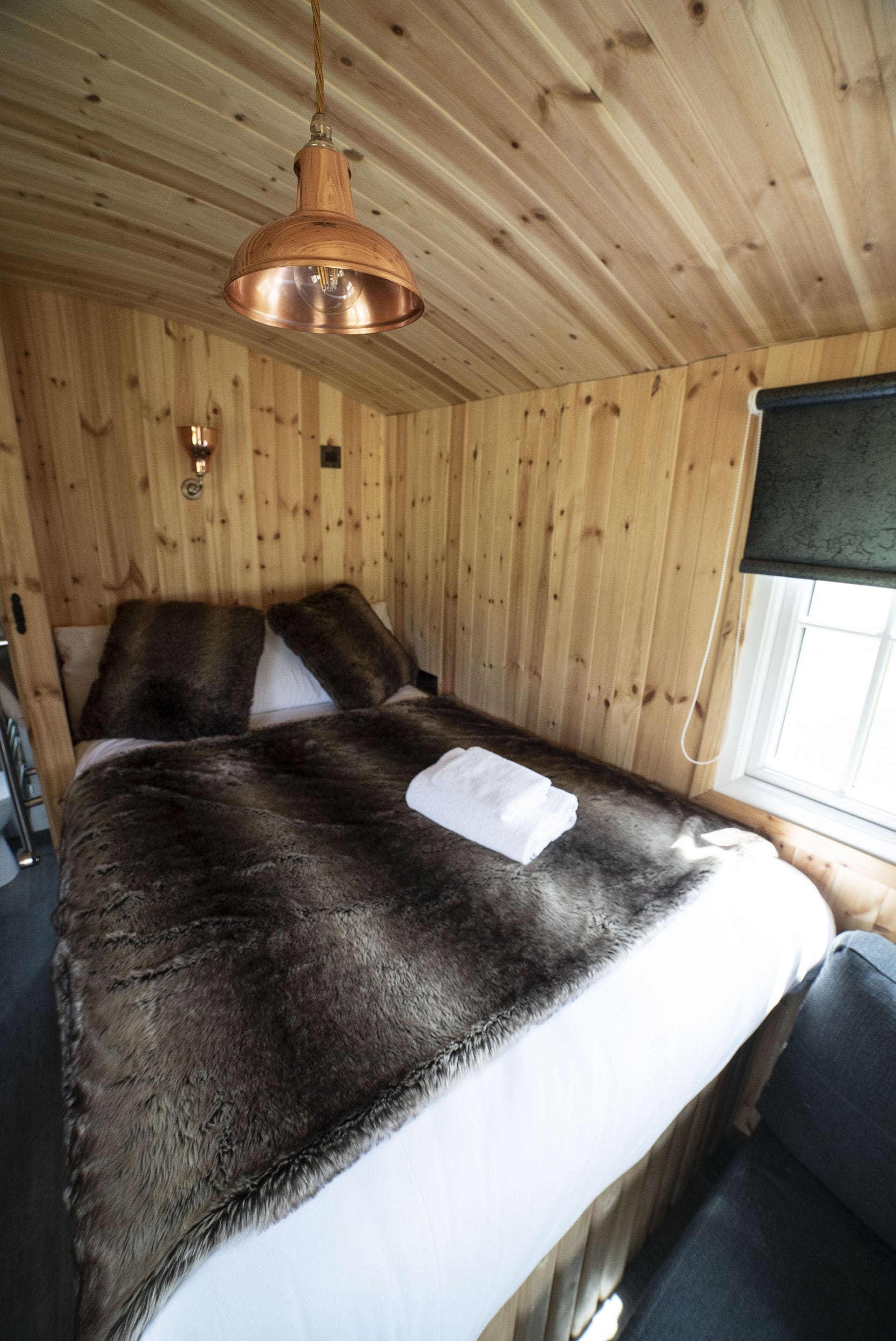 Une pièce de cabane en bois comprenant un lit, un luminaire suspendu et une fenêtre avec un store.