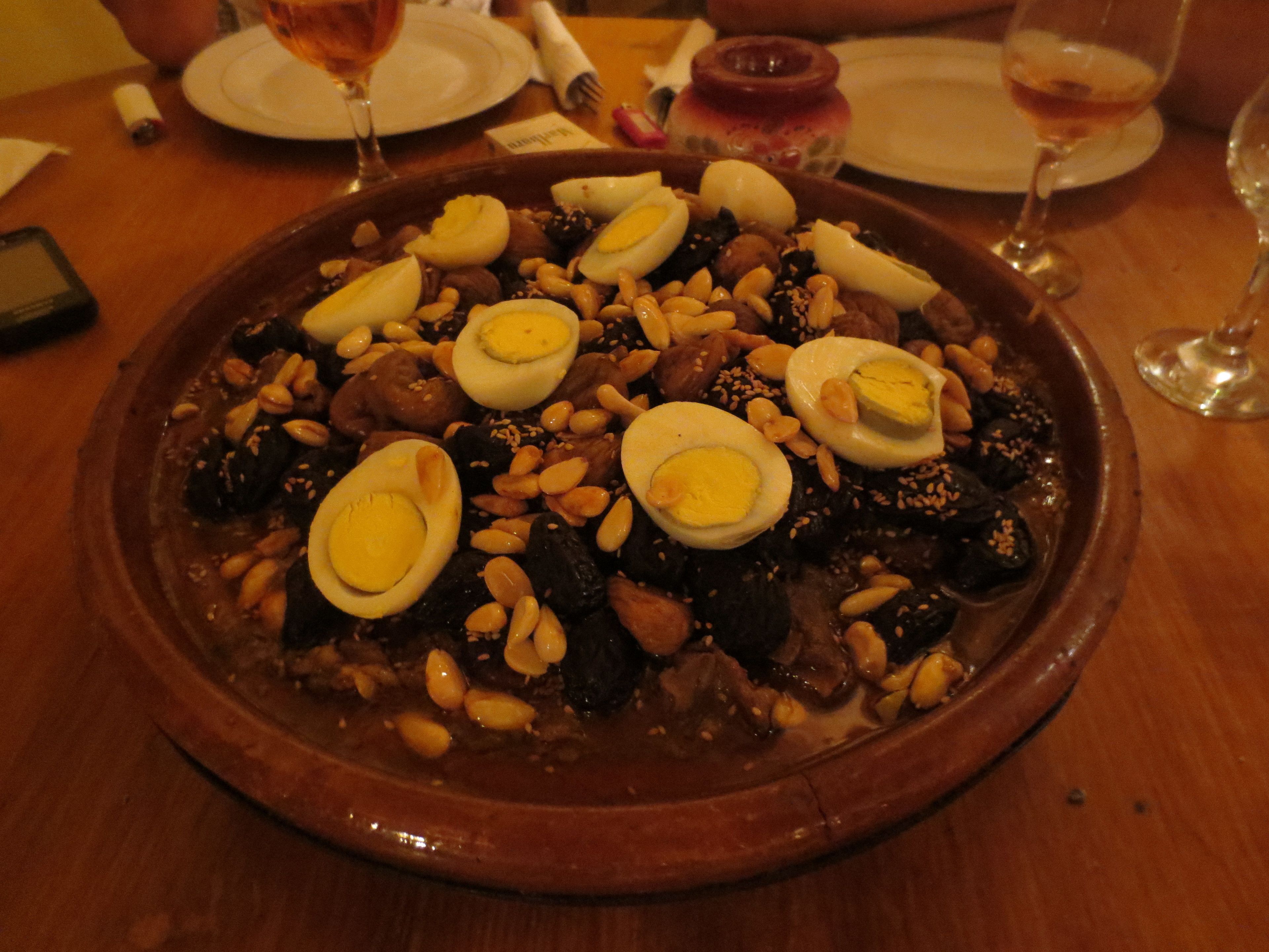 A large, round, brown ceramic dish filled with a variety of foods including nuts, dates, and sliced boiled eggs, placed on a wooden table.