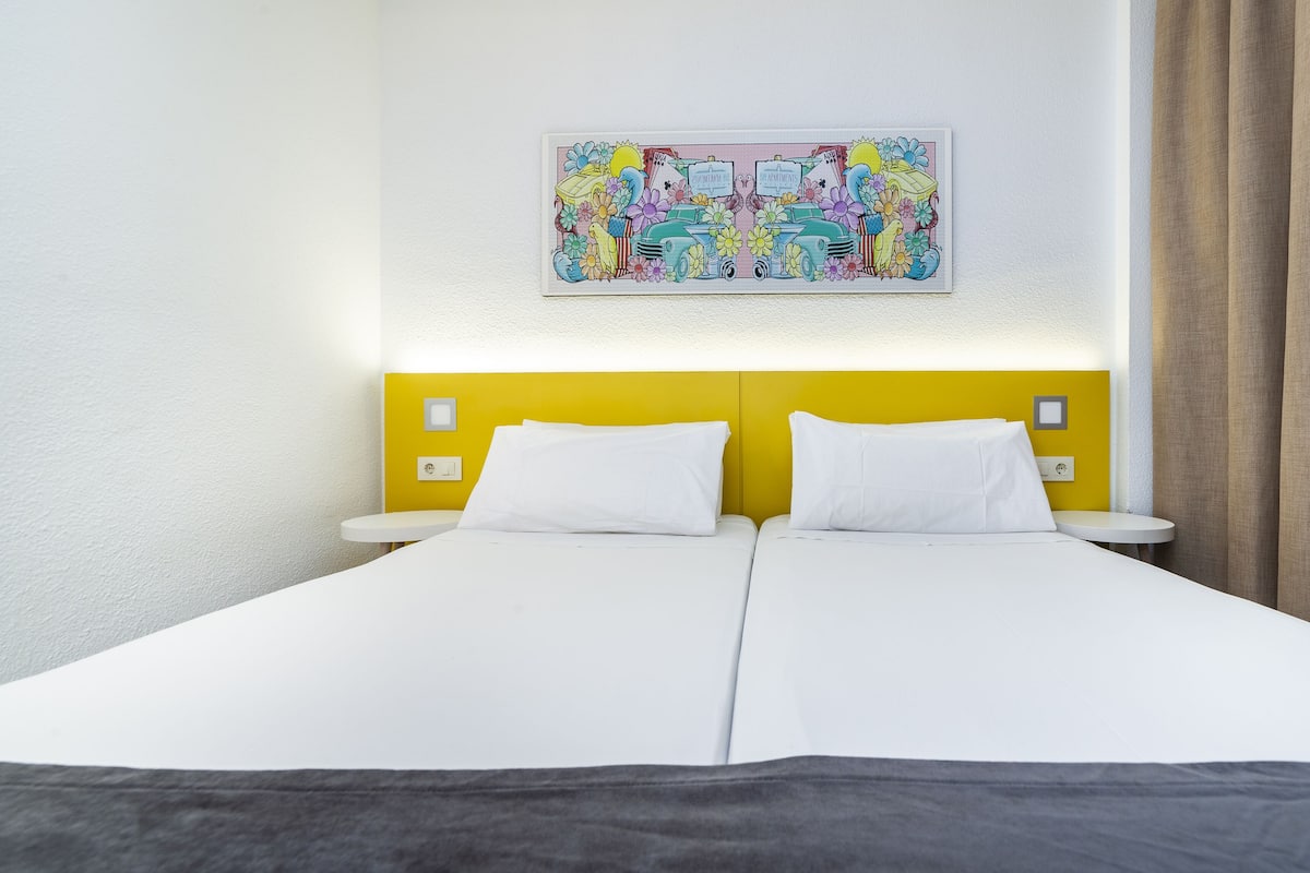 A double bed with white linens, a yellow headboard, and a colorful artwork above it.