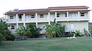 A two-story building with a flat roof, a balcony on the second floor, and a front lawn with tropical plants.