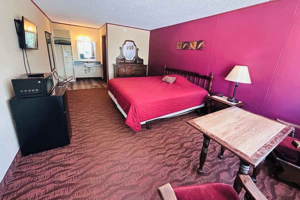 A hotel room with a bed, bedside table, lamp, dresser, mirror, and a desk with a chair.
