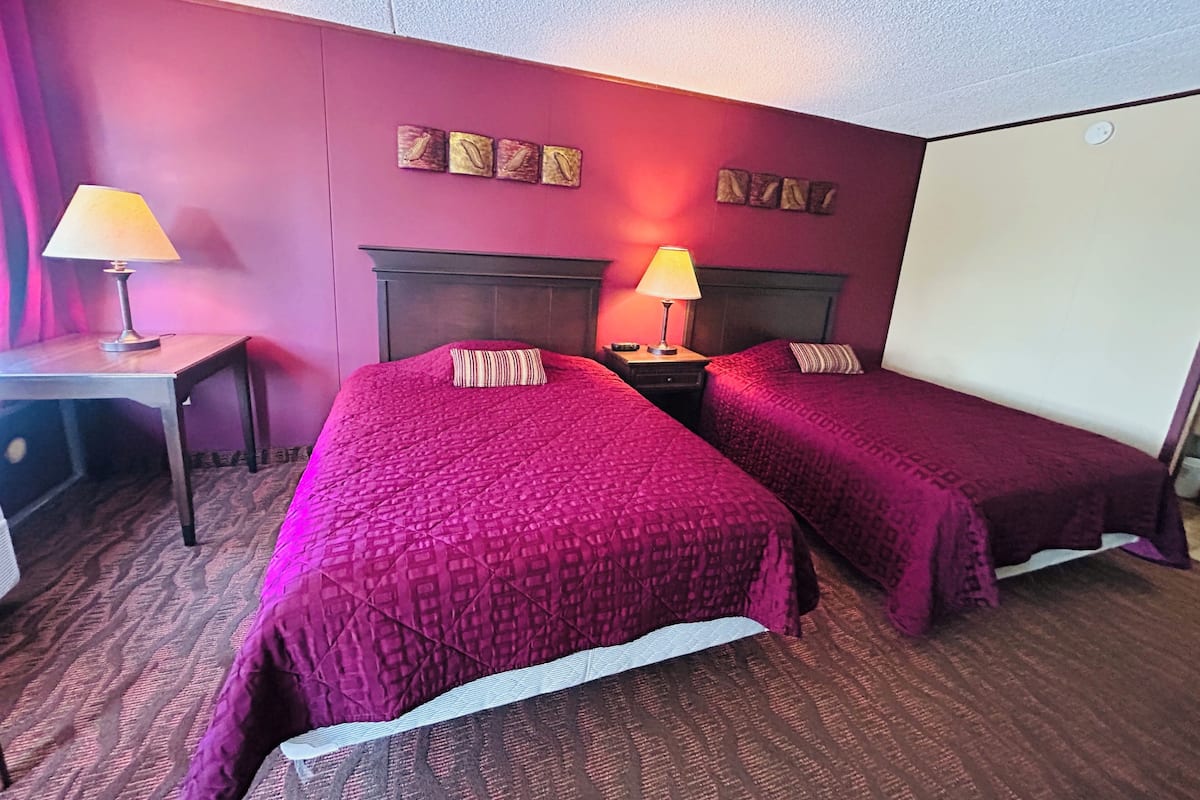 A hotel room with two beds, a desk, a lamp, a nightstand, and a wall-mounted TV.