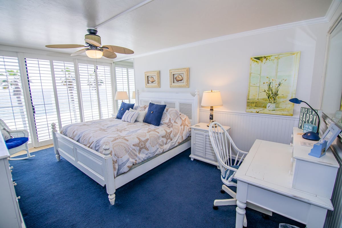 A bedroom with a bed, two nightstands, a desk, a chair, a ceiling fan, and a window with blinds.