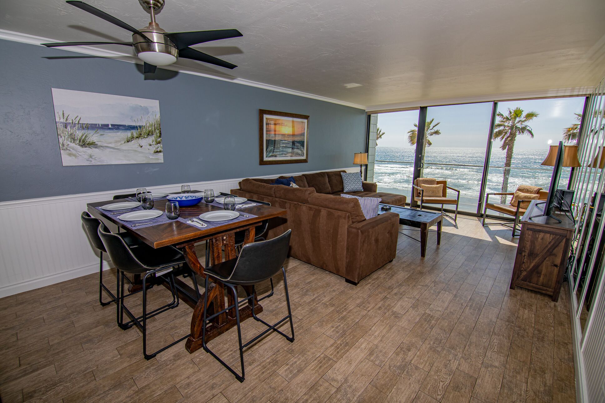 A dining area with a table set for four, a brown sofa, and a view of the ocean through large windows.