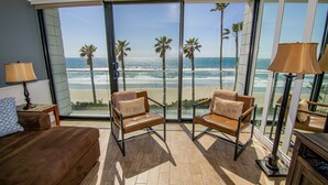 A room with a large window overlooking a beach, two leather chairs with cushions, a brown sofa, and a lamp. - Capri Beach Accommodations (San Diego)