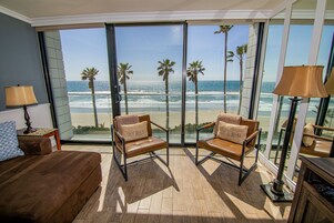 A room with a large window overlooking a beach, two leather chairs with cushions, a brown sofa, and a lamp.