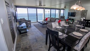 A modern living room with a large window overlooking the ocean, a dining area with a black table set for six, and a gray sofa with red cushions. - Capri Beach Accommodations (San Diego)