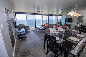 A modern living room with a large window overlooking the ocean, a dining area with a black table set for six, and a gray sofa with red cushions.
