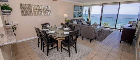 A dining area with a table set for six, a ceiling fan, and a view of the ocean through large windows.